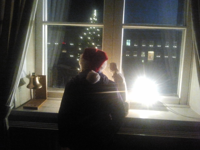 Tomten tittar ut genom fönstret och vinkar till folket som samlats på torget. Alla där nere väntar på luciakröningen.